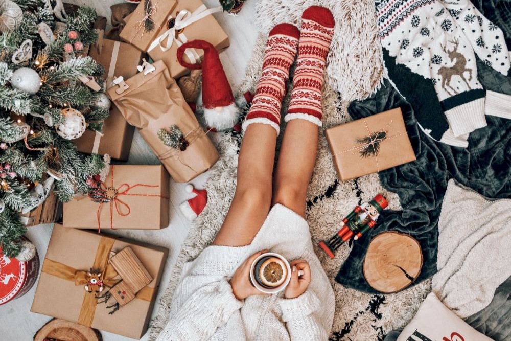 A woman is sitting on the floor next to a Christmas tree and presents.