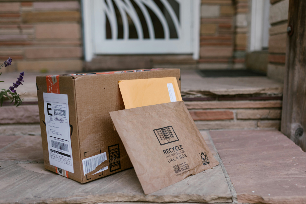 A cardboard box, a brown paper package, and an envelope are placed on a stone doorstep in front of a home's entrance.