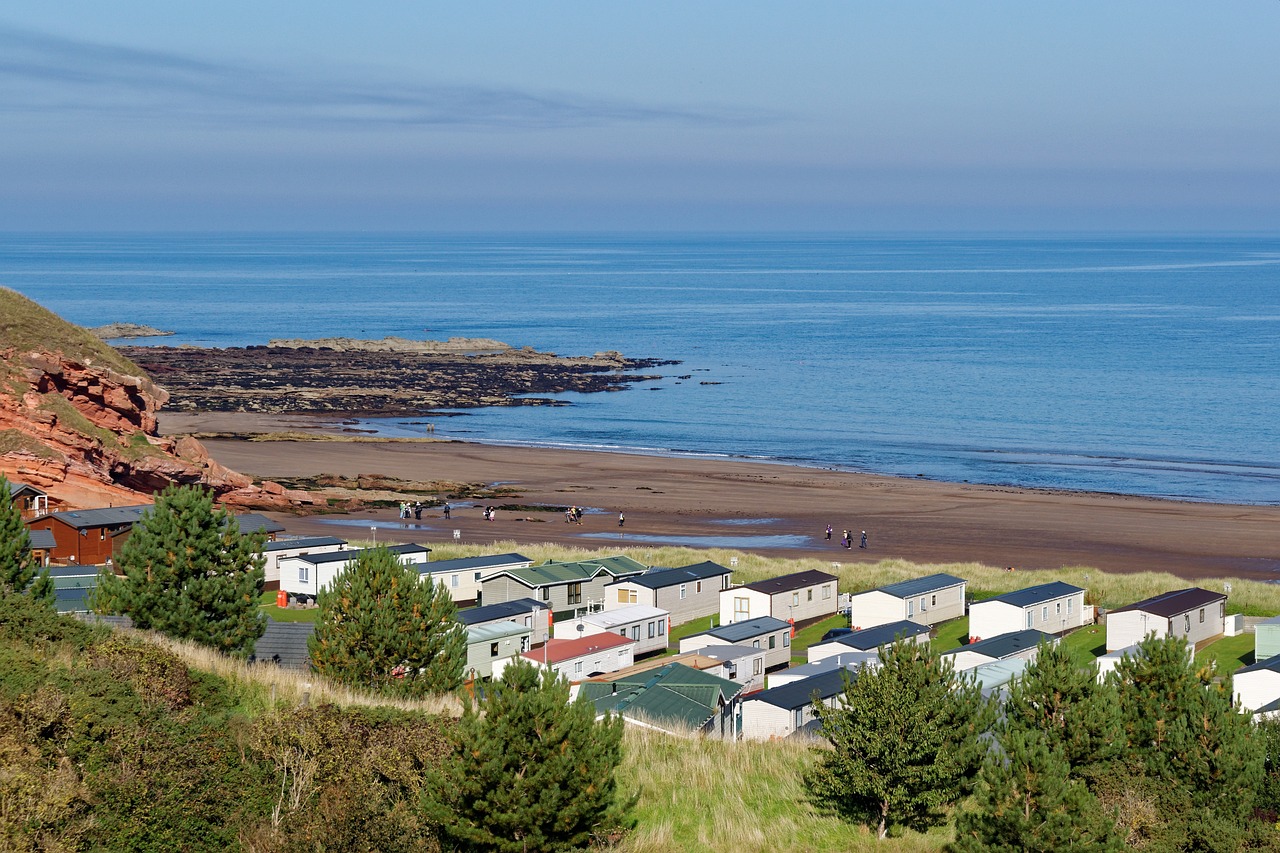 A coastal scene with mobile homes in the foreground, a sandy beach, rocky shoreline, and a calm sea in the background under a clear sky. People can be seen walking on the beach, enjoying their sun holidays 2024.