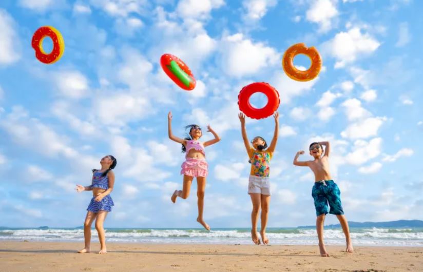 Four children playing on a sandy beach under a blue sky, throwing inflatable rings into the air. The ocean waves are visible in the background—an ideal scene for sun holidays 2024.