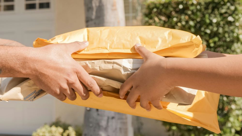 Two hands exchanging a bundle of packages; one hand receiving, the other handing over several yellow and brown envelopes, outdoors with greenery in the background.