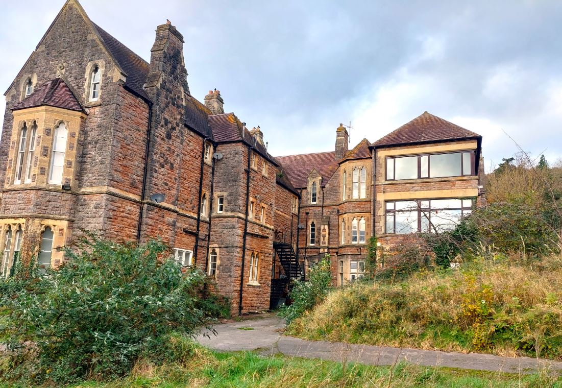 A large, old brick building with tall windows and steep, pitched roofs, surrounded by overgrown greenery and a concrete pathway, offers a unique opportunity for property guardians to save on rent in expensive cities.