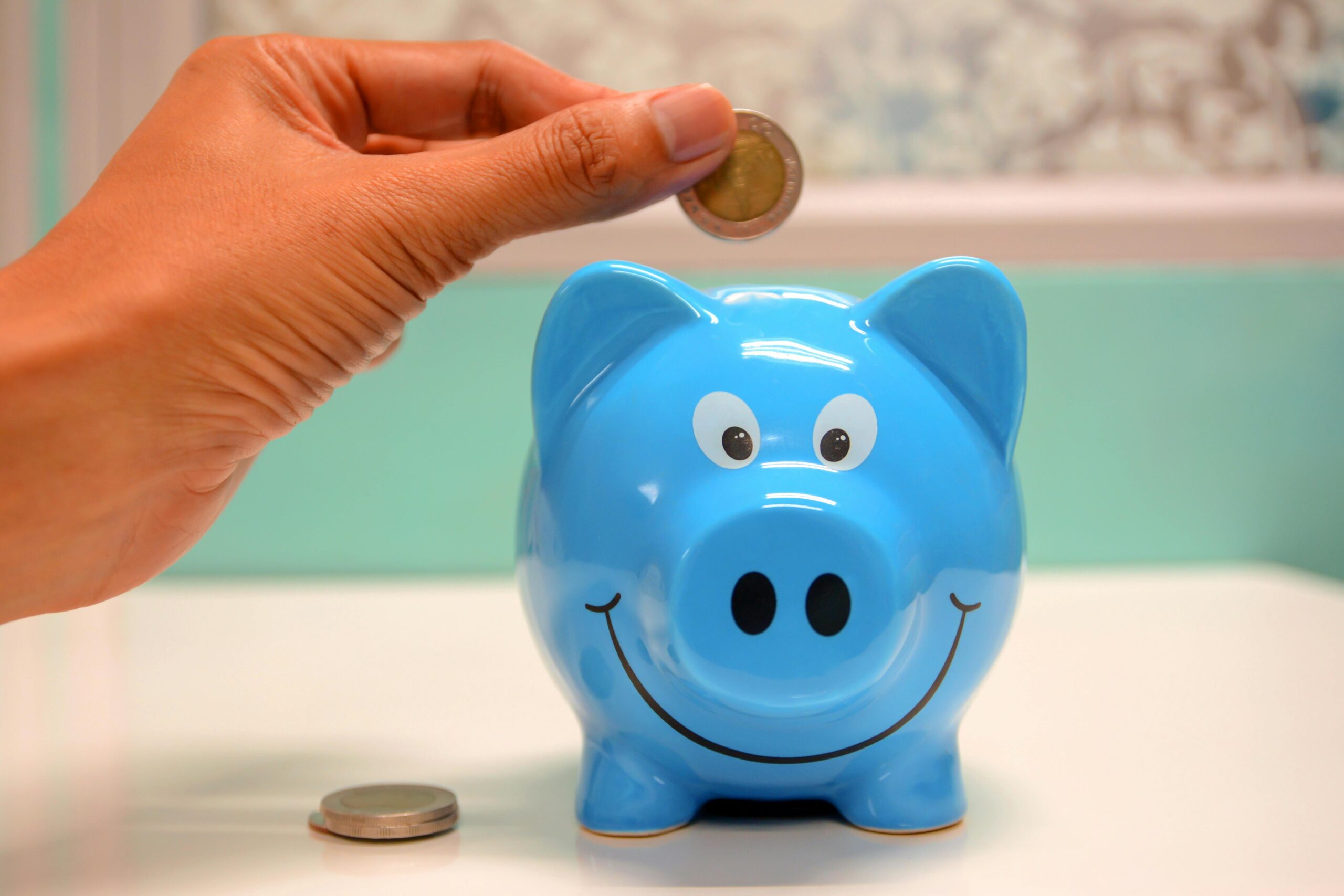 A hand places a coin into a blue piggy bank, which has a smiling face. Two additional coins are on the table nearby.