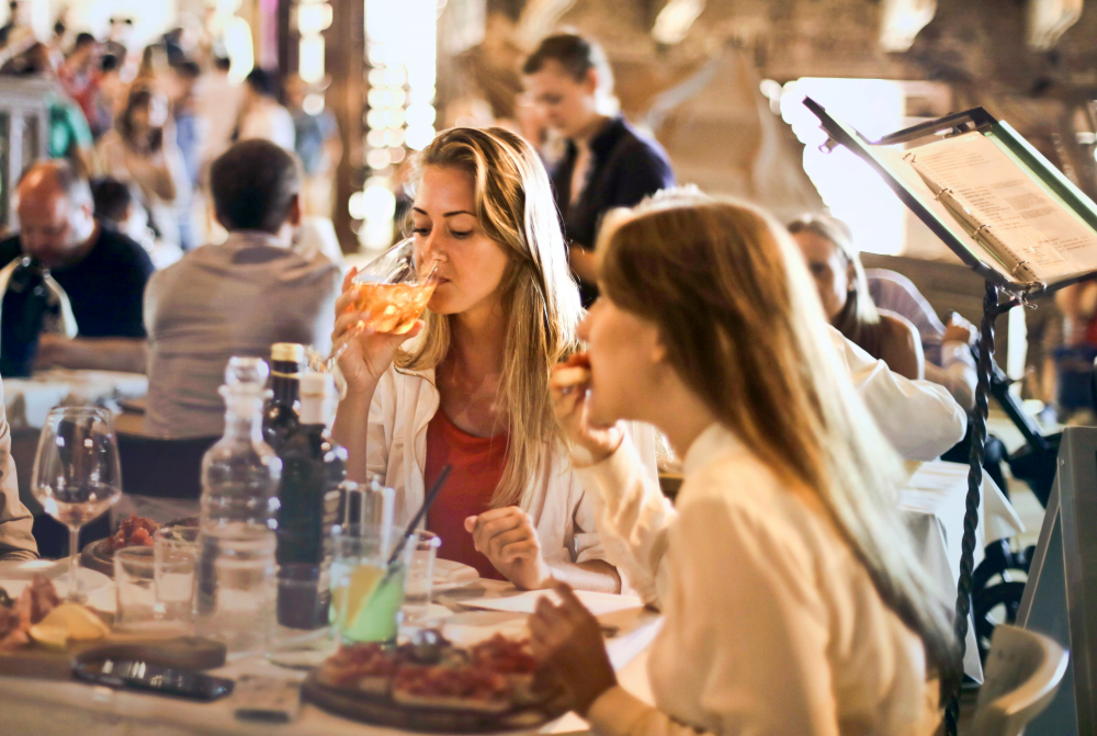 Two women are seated at a restaurant table; one drinks from a glass and the other eats a meal. Other patrons and waitstaff are visible in the background.