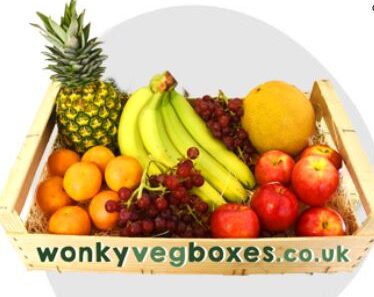 A wooden crate filled with various fruits including a pineapple, bananas, apples, grapes, oranges, and a melon. The crate has the text "wonkyvegboxes.co.uk" on the front.