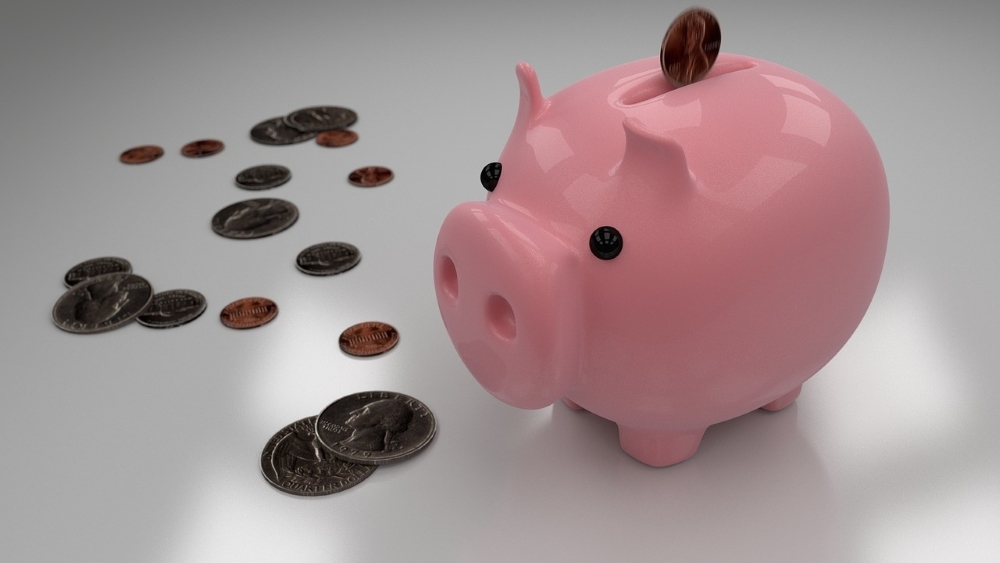 A pink piggy bank with a coin inserted into its slot stands next to scattered coins on a smooth, reflective surface.