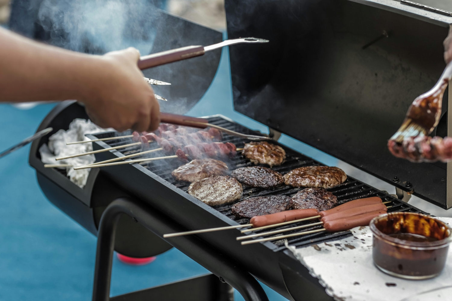 People grilling skewers, burgers, and hot dogs on a barbecue grill with smoke rising and a container of sauce nearby.