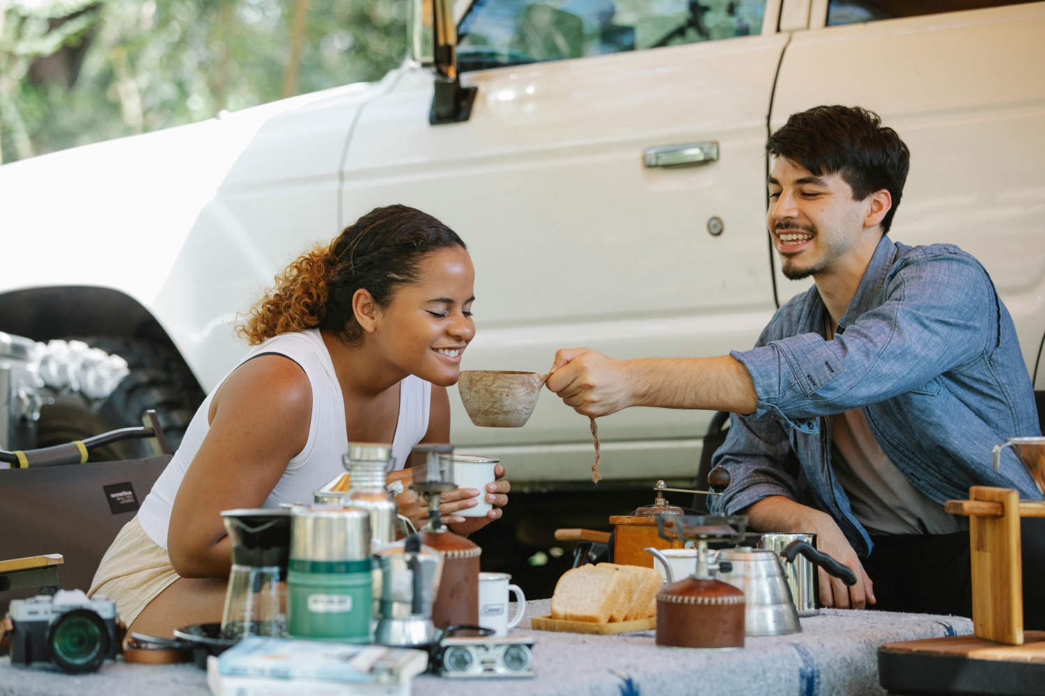 Two people sitting outdoors near a vehicle, with various camping equipment and food items around them. The man is holding a cup towards the woman, who is smiling and leaning in.