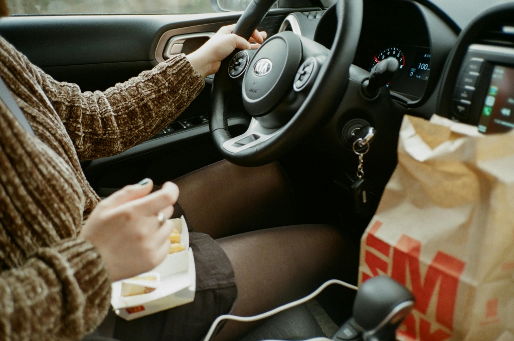 Person eating fast food while driving a Kia car, with a bag of McDonald's on the passenger seat and one hand on the steering wheel.