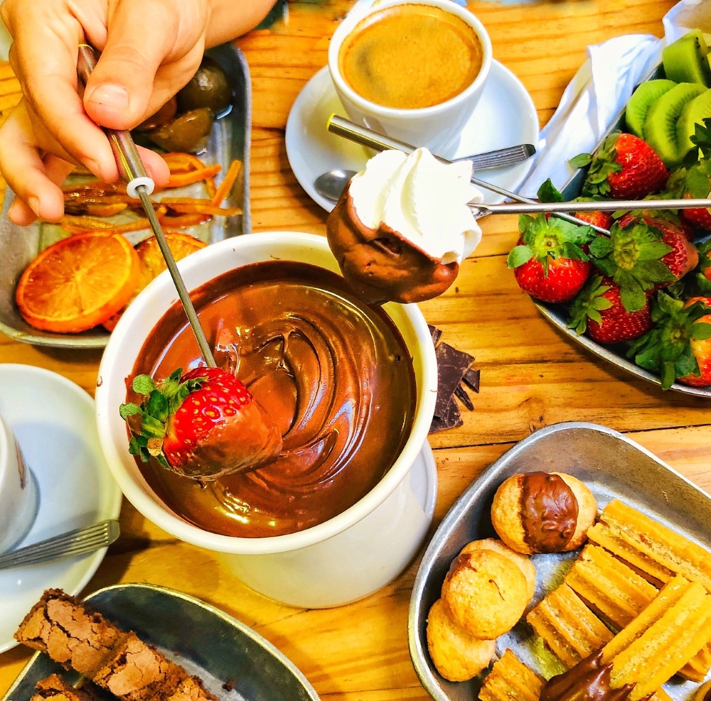 Various fruits and pastries being dipped in a pot of chocolate fondue, accompanied by a cup of coffee on a wooden table.