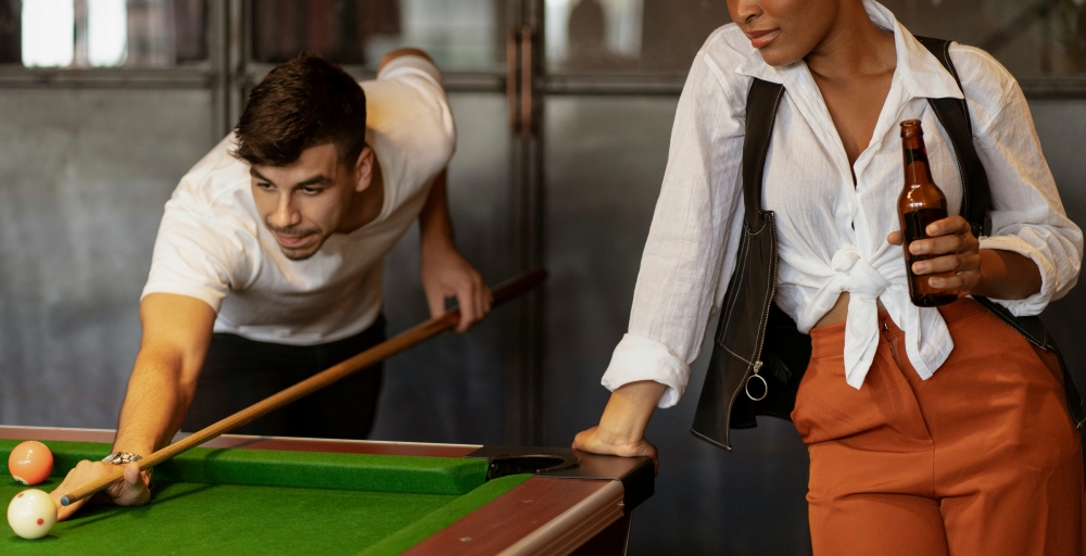 Two people are at a pool table; one is aiming a cue stick to take a shot, while the other leans on the table holding a beer bottle.