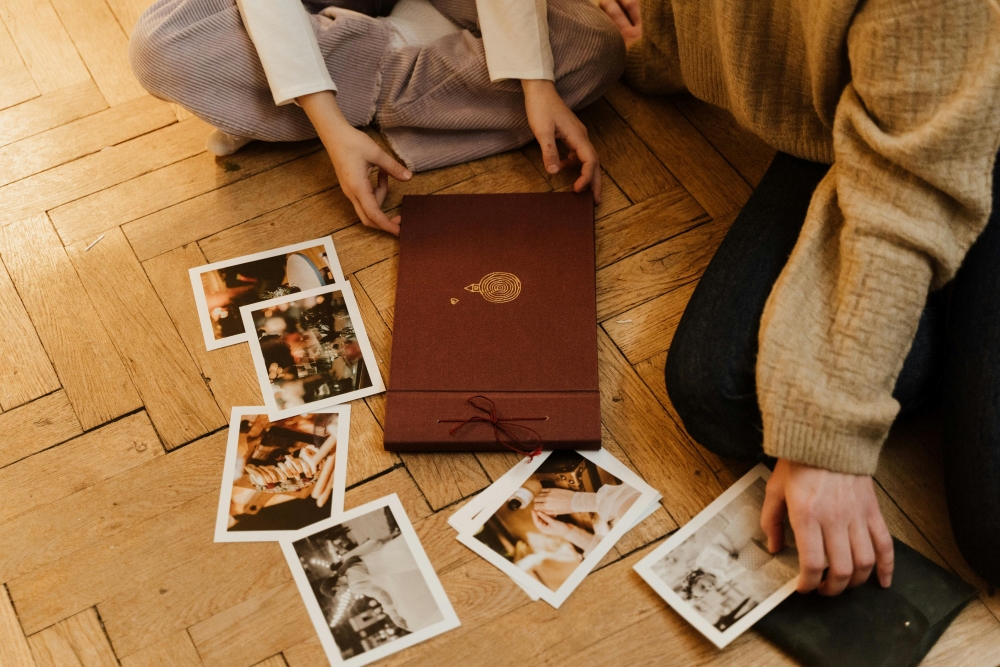 Two individuals sit on a wooden floor, looking through a red photo album. Several loose photographs are spread out around the album.