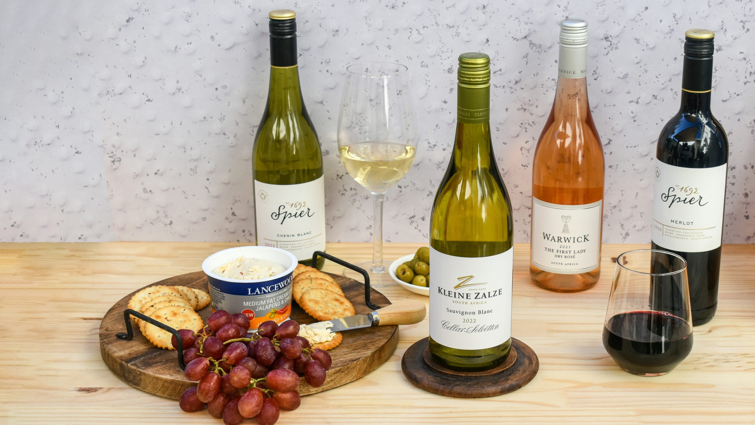 A selection of four wine bottles, two glasses of wine, a bowl of cheese, grapes, crackers, and green olives arranged on a wooden surface.