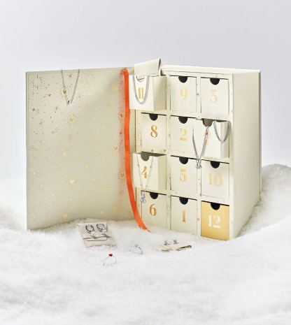 An advent calendar with small drawers numbered 1-12, containing jewelry items such as necklaces, bracelets, rings, and earrings, displayed on a white fluffy surface.