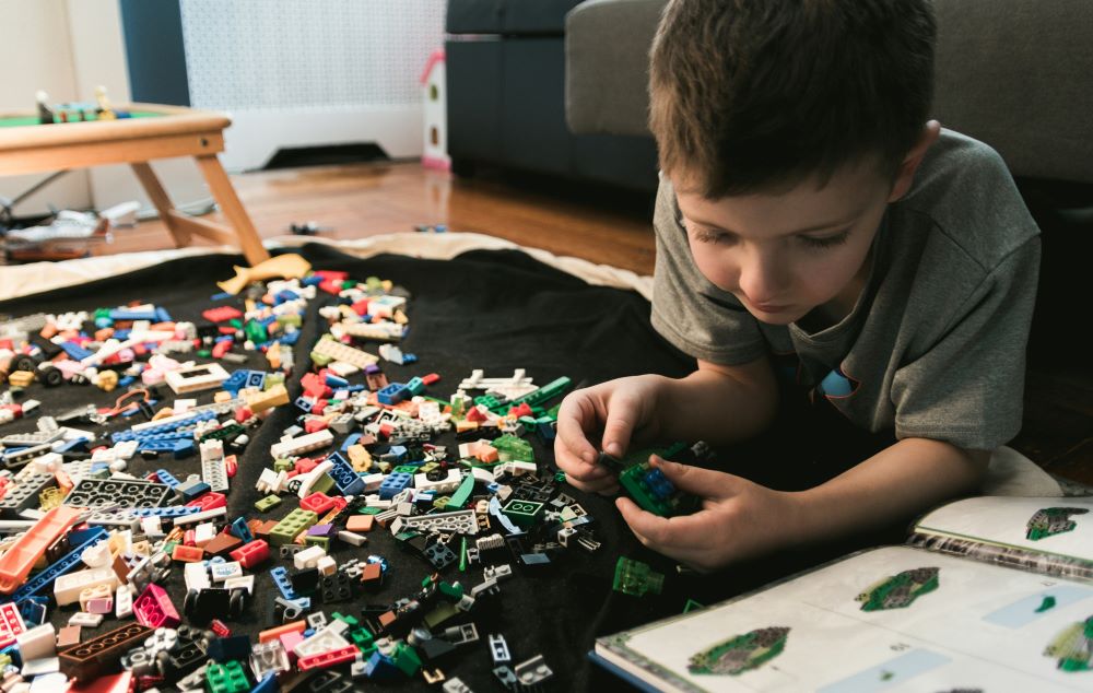 A child lying on the floor, focused on building with colorful Lego blocks scattered around, with an instruction booklet next to them.