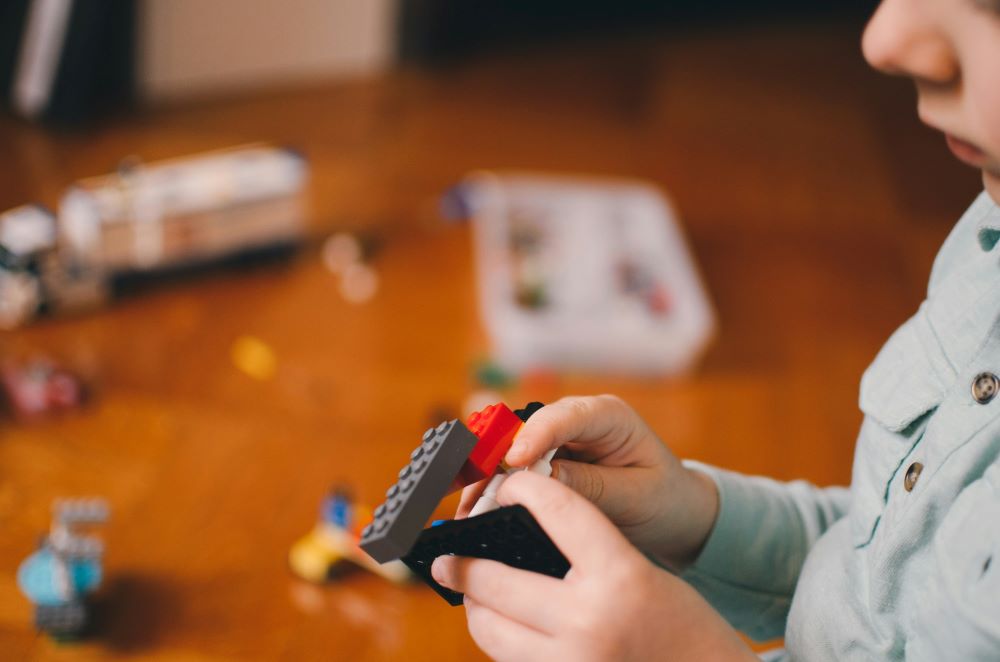 Child playing with plastic building blocks on a brown surface, with more blocks and parts scattered in the background.