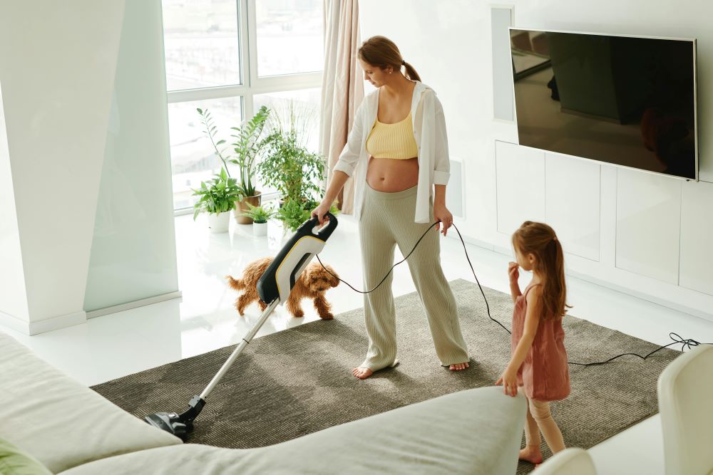 A woman vacuums a living room rug near a large window. A small child stands nearby, and a dog jumps onto the rug.