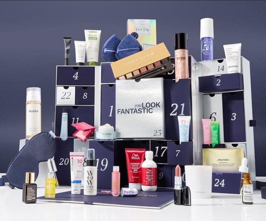 A display of various skincare and beauty products arranged in a calendar-like structure with numbered compartments, featuring items such as creams, serums, and makeup products on a blue background.