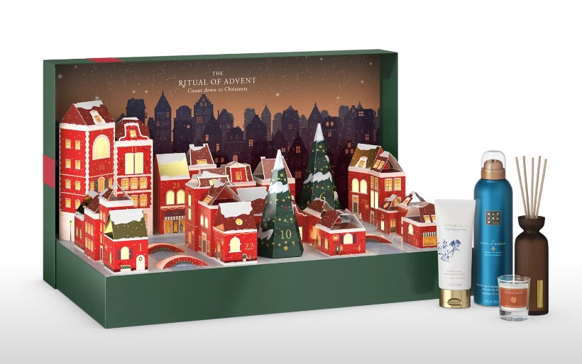 A festive-themed advent calendar with various small houses and a tree, accompanied by skincare and home fragrance products, including a bottle, spray, and a reed diffuser.