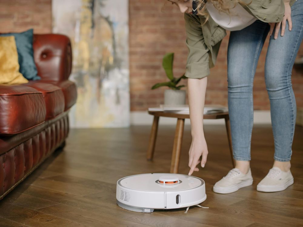 A person is about to press a button on a white robot vacuum cleaner on a wooden floor, with a red couch and a wooden table in the background.