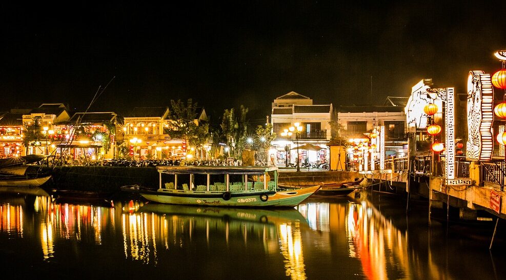 A night view of a riverside town with illuminated buildings, colorful lanterns, boats on the water, and reflections on the river.