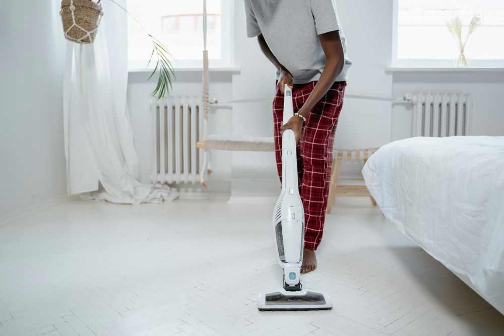 A person in plaid pajama pants uses a cordless vacuum to clean a white bedroom floor with minimalistic decor and natural light from two windows.