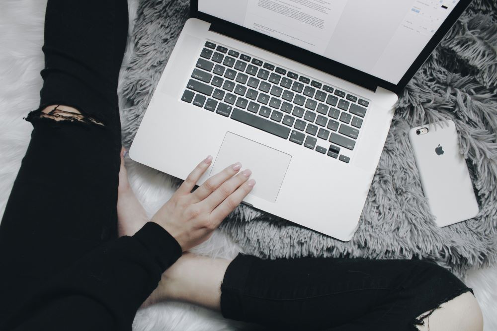 Person using a laptop on a gray furry surface with an iPhone beside them.