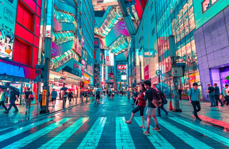 A busy street in a city with colorful, illuminated buildings, people crossing a zebra crossing, and signs in Japanese.