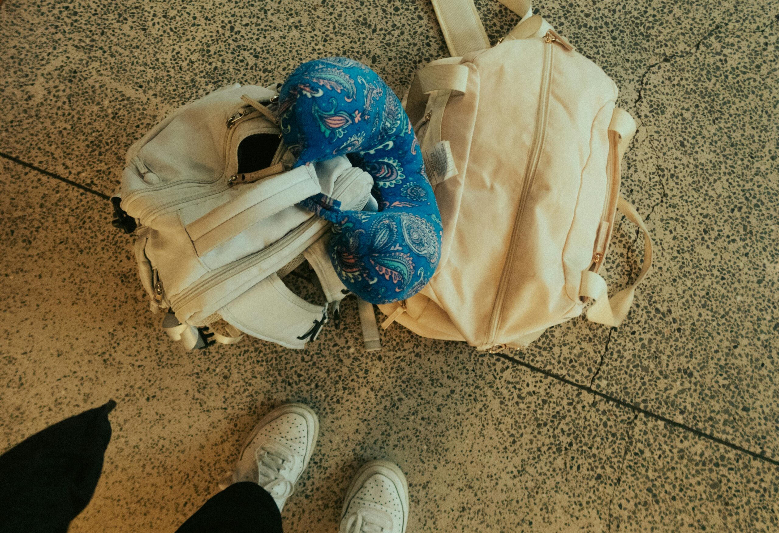 Two white bags and a blue patterned neck pillow are on a speckled floor. The feet of a person wearing white sneakers are partially visible at the bottom of the image.