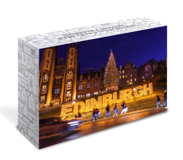 A lighted "EDINBURGH" sign and a large Christmas tree are illuminated at night with pedestrians passing by.