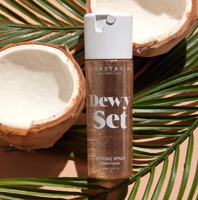 A bottle of Anastasia Dewy Set setting spray is placed on palm leaves, next to two coconut halves.