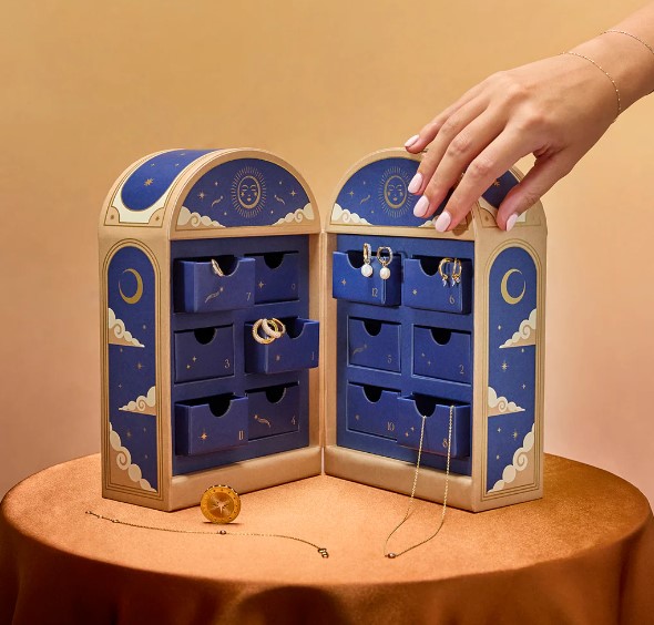 A hand reaches to open a drawer of a jewelry advent calendar with blue and gold designs, displaying various pieces on each side, set on a round table with a few items placed beside it.