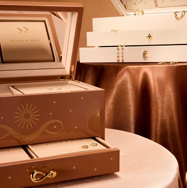 A brown jewelry box with star designs is open, revealing organized compartments. Nearby, white jewelry boxes are stacked on a satin-covered table.