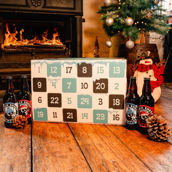 Advent calendar box on a wooden floor with four beer bottles and pine cones. A fireplace and Christmas tree are in the background.