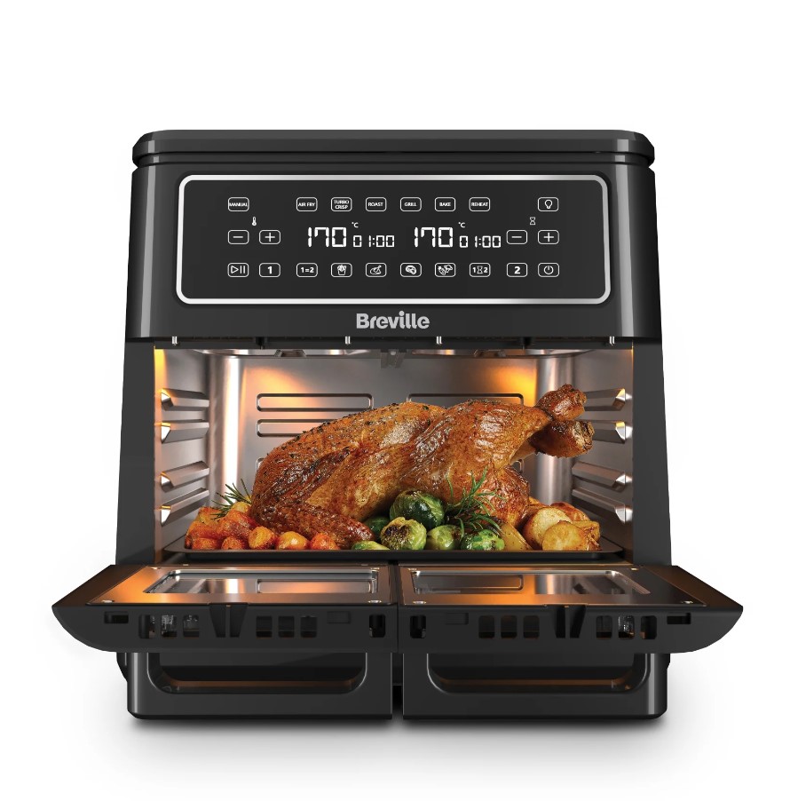 A black Breville oven with digital controls, cooking a large roast chicken accompanied by vegetables inside. The oven door is open, revealing the food.