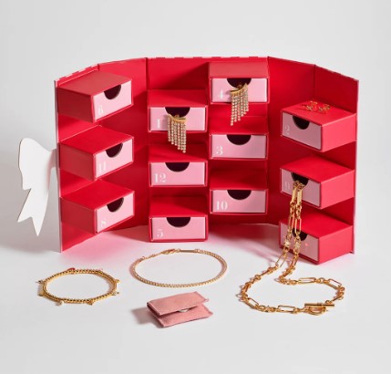 A red and pink advent calendar box with several drawers open, revealing gold jewelry including necklaces and bracelets displayed on a surface.