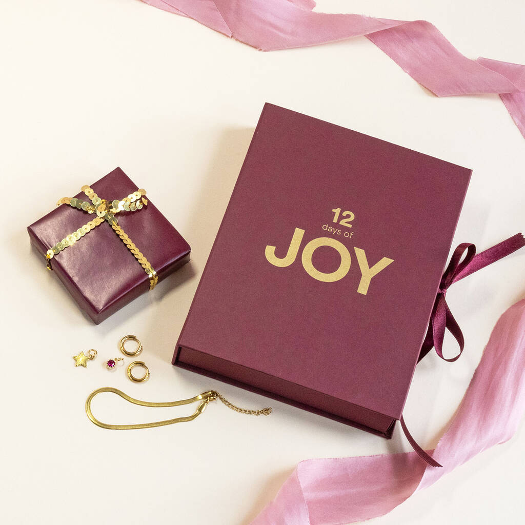 Gift set titled "12 days of JOY" with purple boxes, one small and one large, surrounded by a golden bracelet, earrings, and pink ribbon.