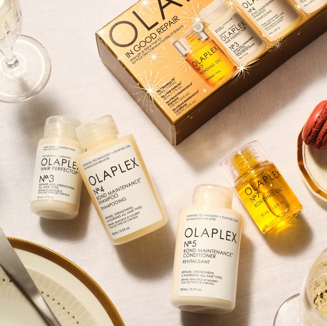 A set of Olaplex hair care products on a table, including shampoo, conditioner, and hair perfector, with a festive gift box and a red macaron nearby.