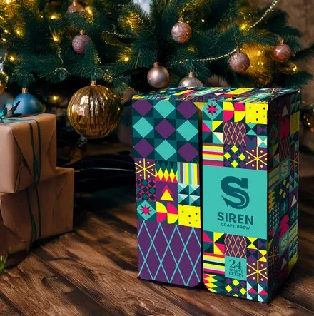 Gift box with colorful geometric patterns, labeled "Siren Craft Brew," on a wooden floor next to a Christmas tree with lights and ornaments.