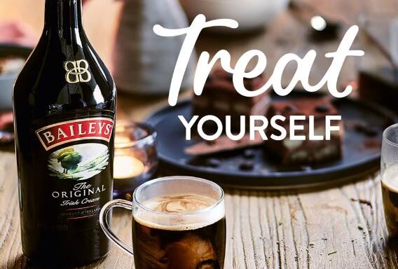 A bottle of Baileys next to a mug of coffee and desserts on a wooden table tempts you with the text "Treat Yourself." Discover the cheapest Baileys 1 litre today and indulge in this delightful offer.