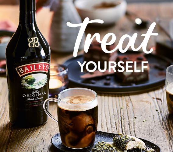 A bottle of Baileys next to a mug of coffee and desserts on a wooden table tempts you with the text "Treat Yourself." Discover the cheapest Baileys 1 litre today and indulge in this delightful offer.