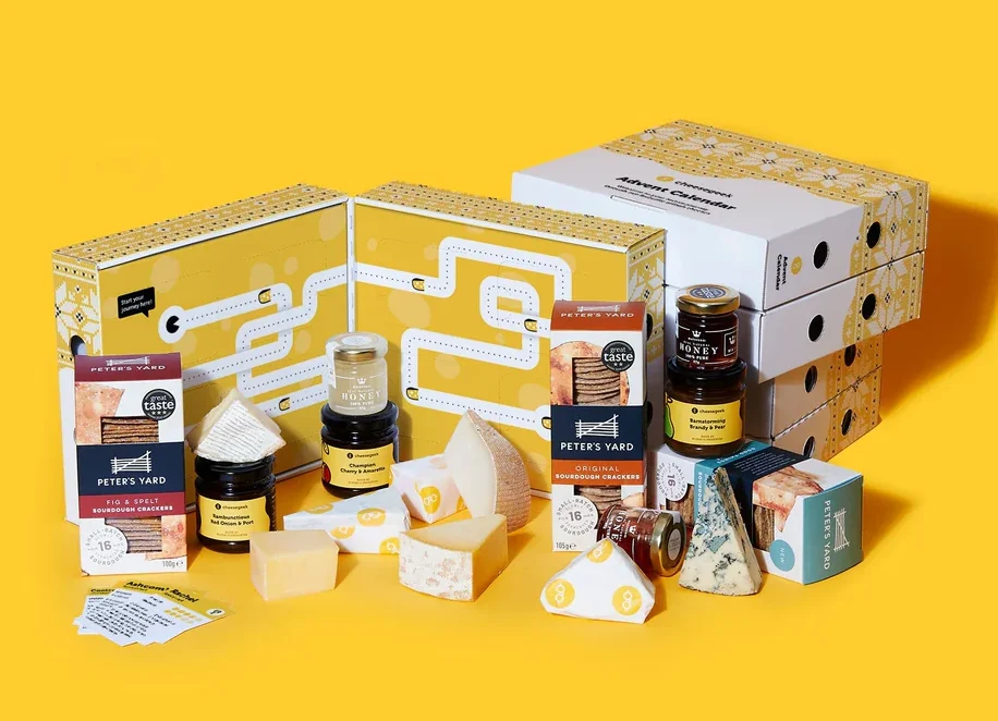 Assorted cheeses, jams, and packaged items are displayed in front of yellow boxes against a yellow background.