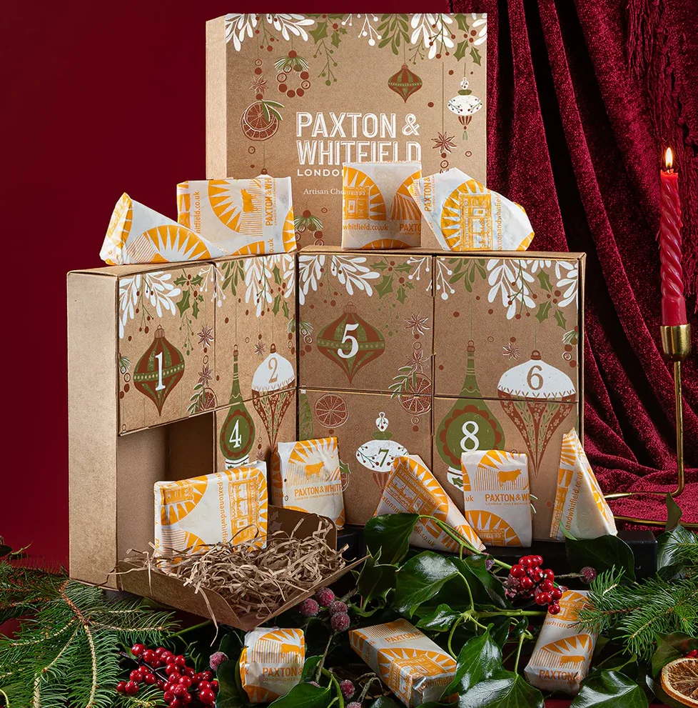 Cheese advent calendar with numbered doors, wrapped cheeses, foliage, berries, and a red candle on a red background.