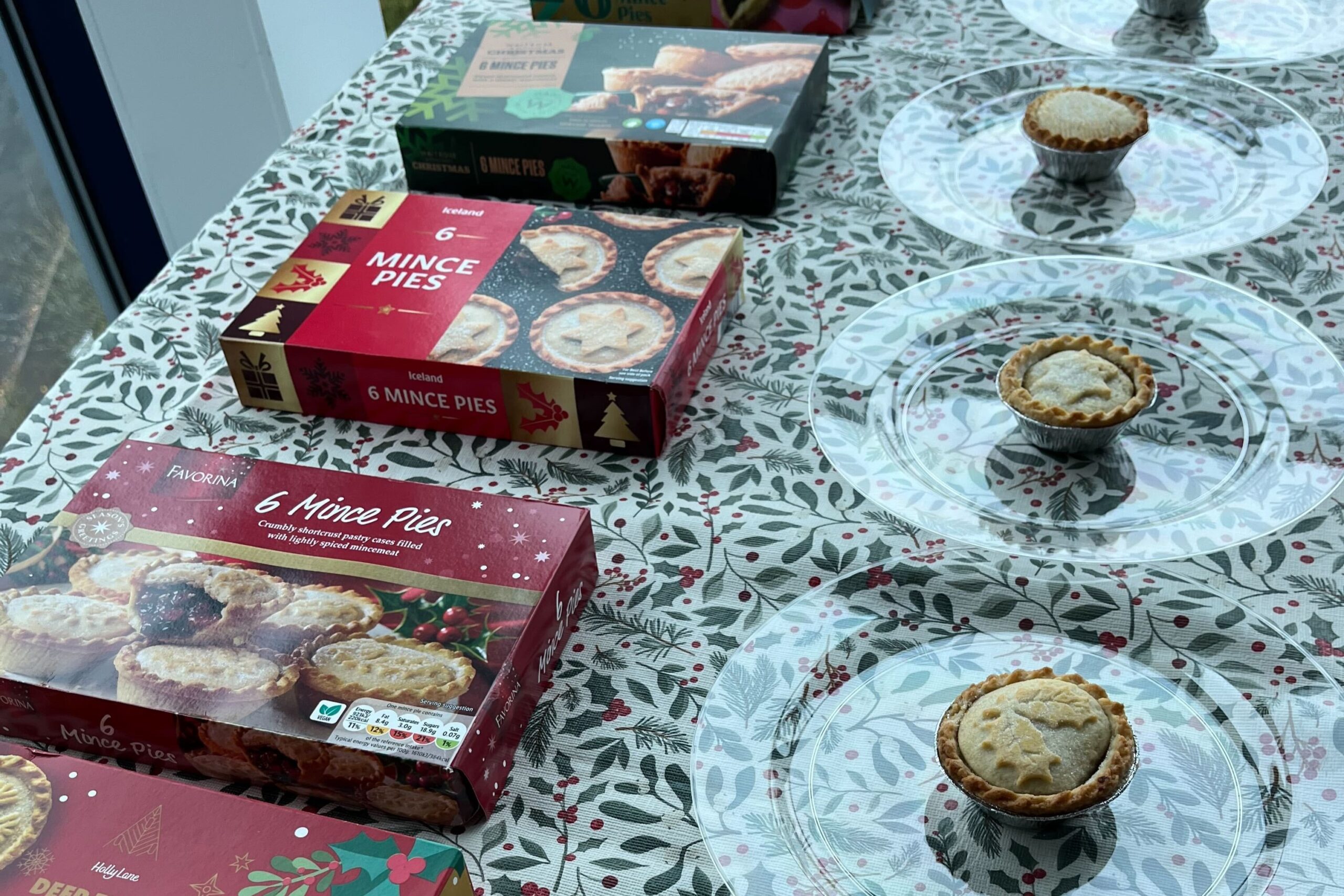 Boxes of supermarket mince pies and individual pies are displayed on clear plates atop a floral-patterned tablecloth.