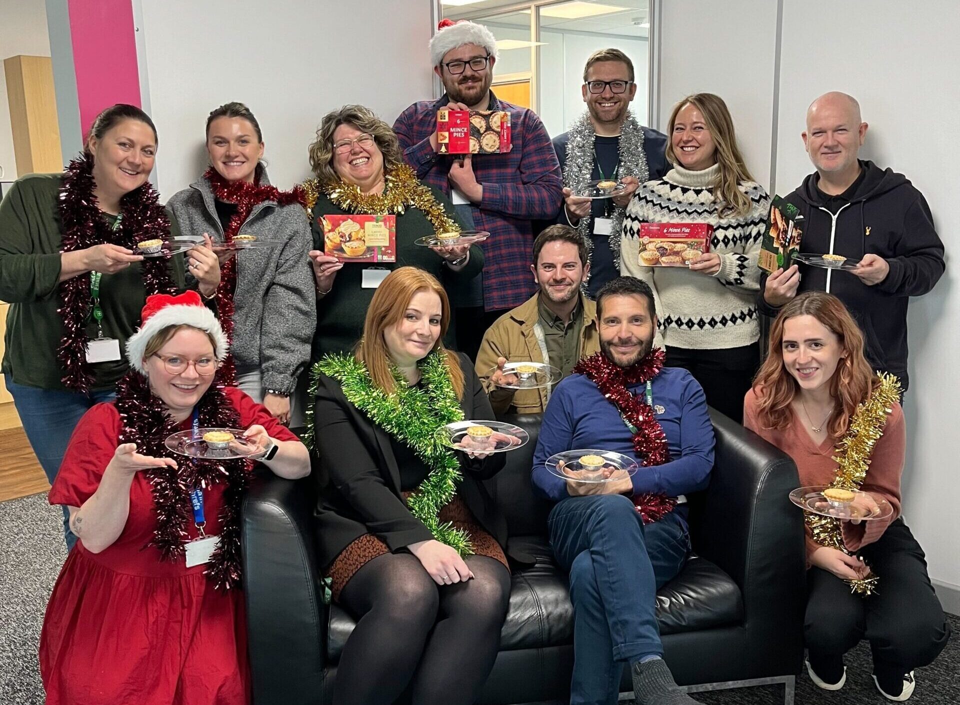 A group of people wearing festive attire gather in an office, holding holiday-themed items. Some wear tinsel and Santa hats while posing for a group photo.