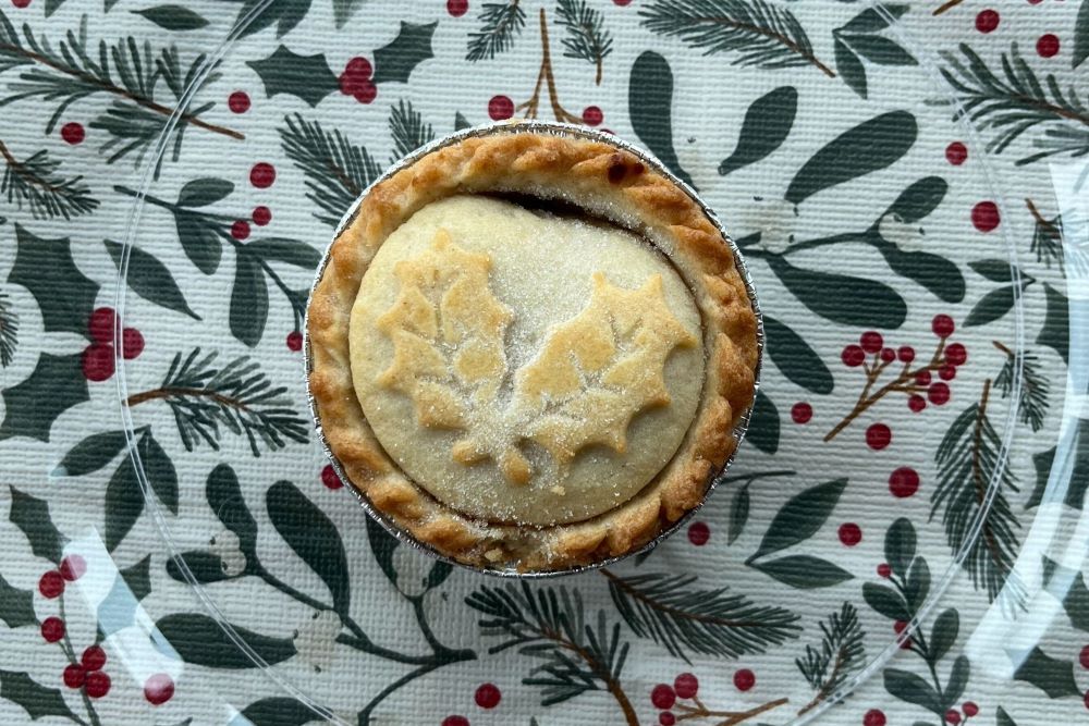 A small pie with a sugared top and holly leaf design sits on a festive tablecloth featuring pine branches and red berries.