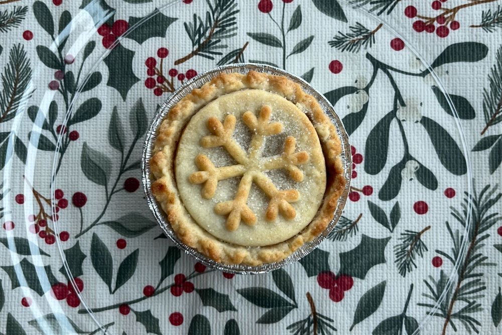 A mince pie with a snowflake-shaped pastry top sits on a clear plate, placed on a tablecloth with a festive holly and berry pattern.