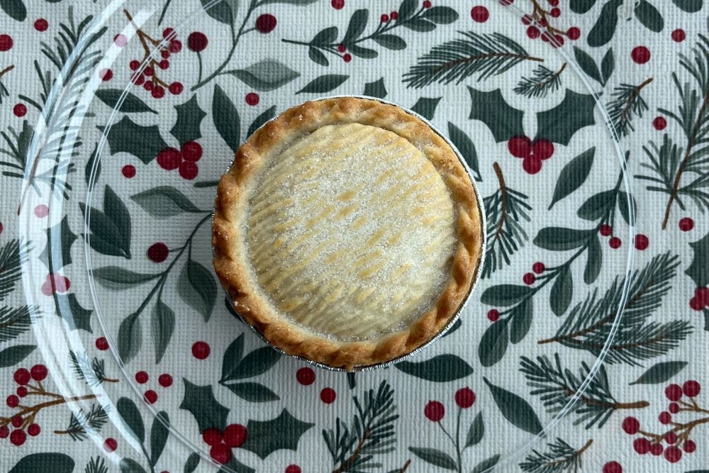 A pie on a clear glass plate sits on a tablecloth with red berries and green pine leaves.