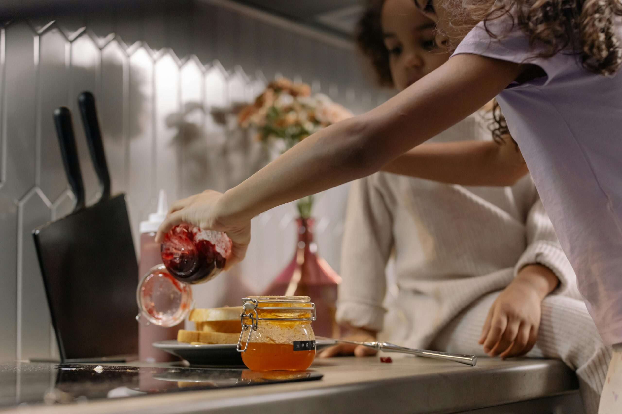 Two children are in a kitchen. One is pouring jam from a jar onto a plate with bread, while the other watches. A knife and an open jar of orange spread are on the counter.