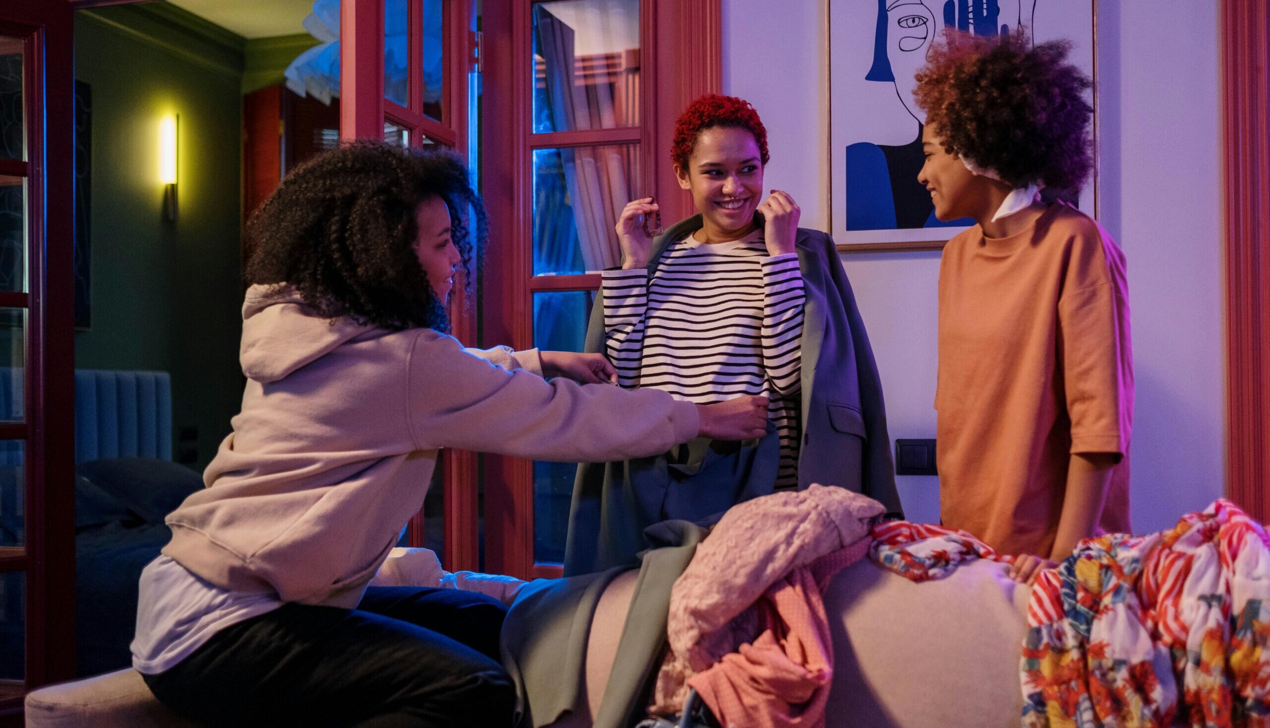 Three people in a room with colorful clothes. One is holding up a striped shirt while the others observe. A painting is on the wall behind them.